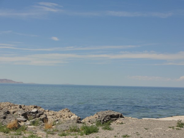 This photo shows a large blue lake with a rocky shore under a blue sky with white clouds, inspiring a #FridayPhotoPoetry post about the best way to travel.